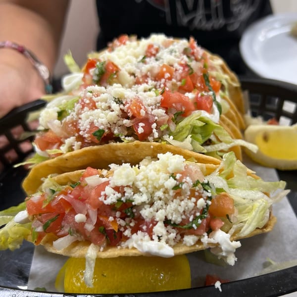 Three Mi Linda Tierra Taqueria tacos with lettuce, pico de gallo, cheese, and a lemon wedge.
