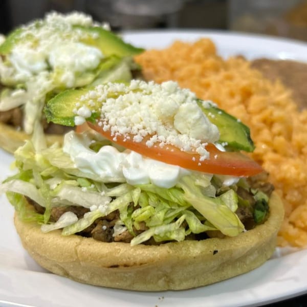 Two Mi Linda Tierra sopes topped with meat, lettuce, sour cream, avocado, tomato, and cheese, with rice and beans.