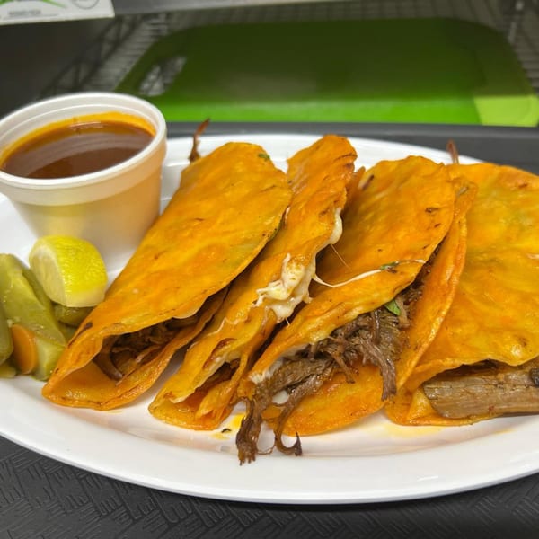 Four birria tacos with melted cheese, a side of consommé, pickled vegetables, and a lime wedge.